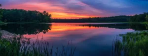 a tranquil sunset casts brilliant hues of orange and pink over a serene lake, reflecting the vibrant sky and surrounded by lush greenery.